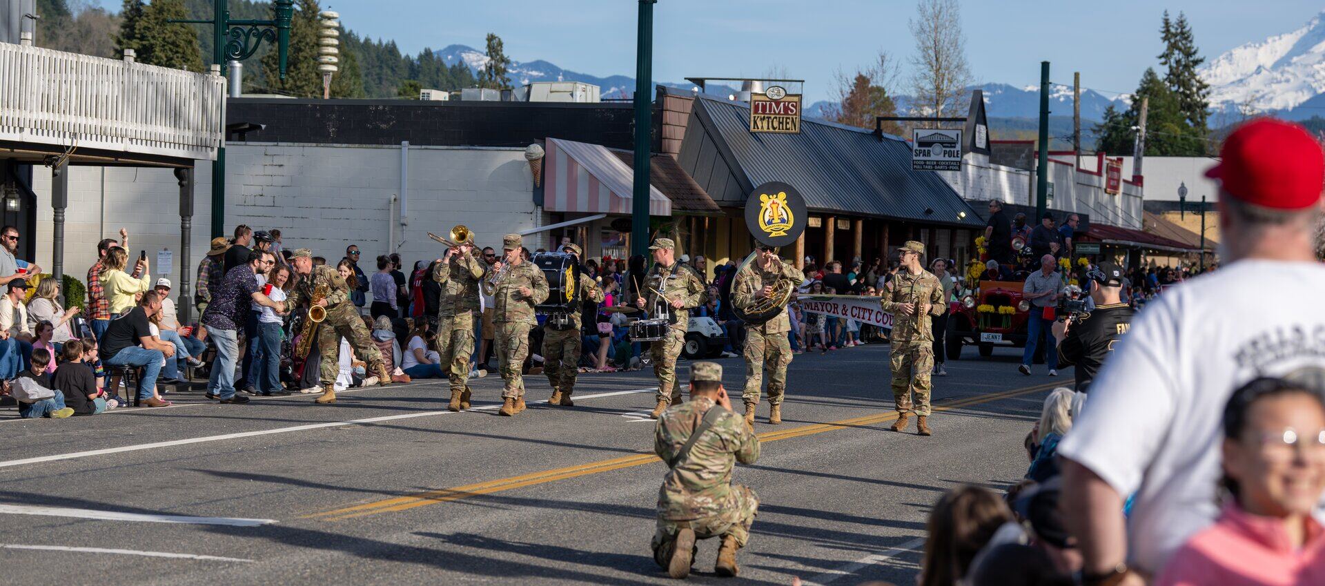 An army band marches past crowds on parade day in downtown Orting, with mountains behind